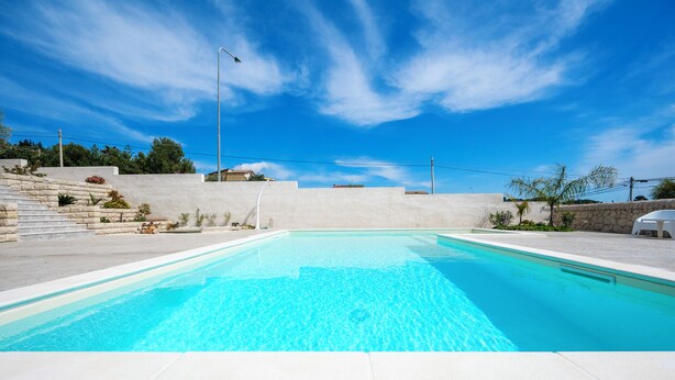 Una piscina con acqua cristallina e blu, circondata da un solarium in piastrelle bianche e un muro in pietra.