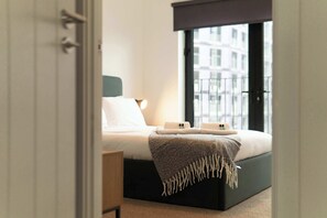 Ein modernes Schlafzimmer mit Bett, Nachttischen und Blick auf ein Gebäude durch ein Fenster.