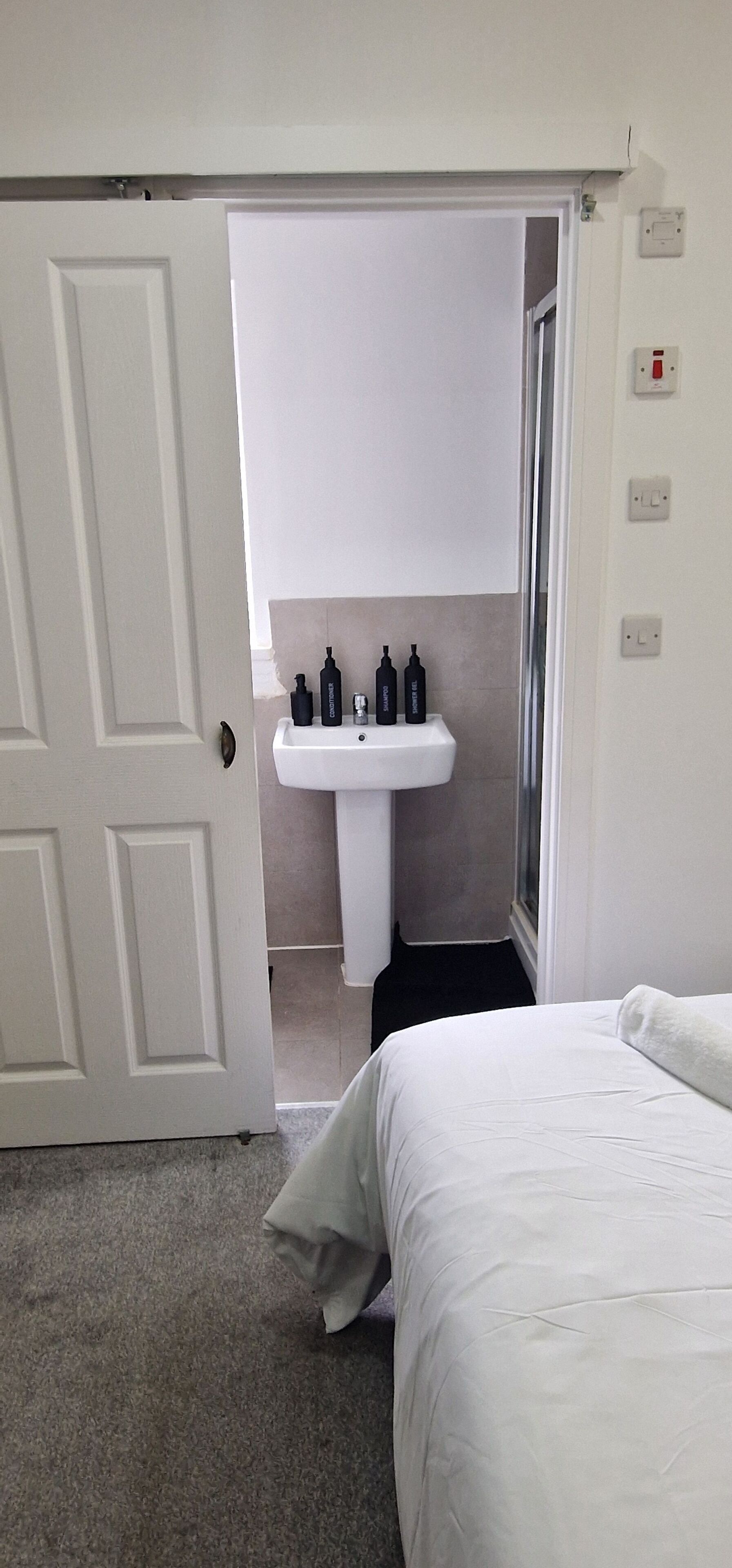 Une chambre avec un lit blanc, une porte blanche, une salle de bain avec un lavabo et des articles de toilette, et une douche.