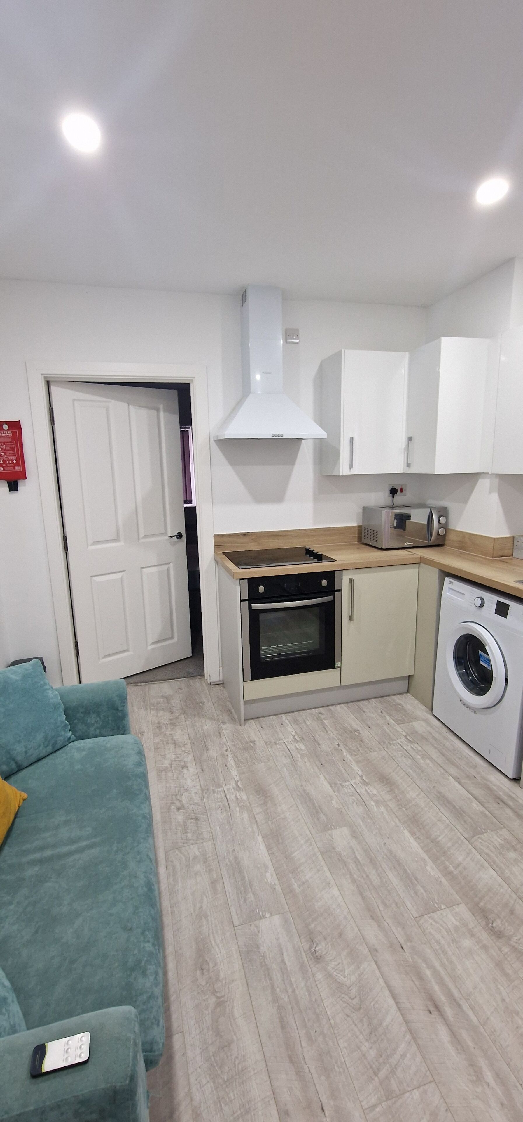 A compact kitchen with a built-in oven, microwave, and washing machine, a teal sofa, and a white door.