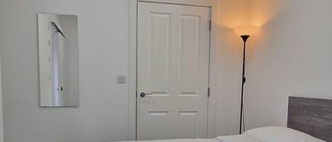 Un lit bien fait, avec une couette blanche et une serviette enroulée, une porte blanche, un miroir et une lampe sur pied dans une pièce dont le sol est recouvert d’une moquette.