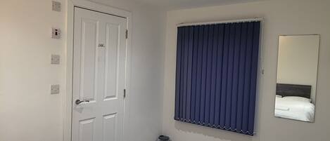 Ett hotellrum med en stor säng, en dörr med handtag, ett fönster med persienner, en liten papperskorg och en spegel som är monterad på väggen.