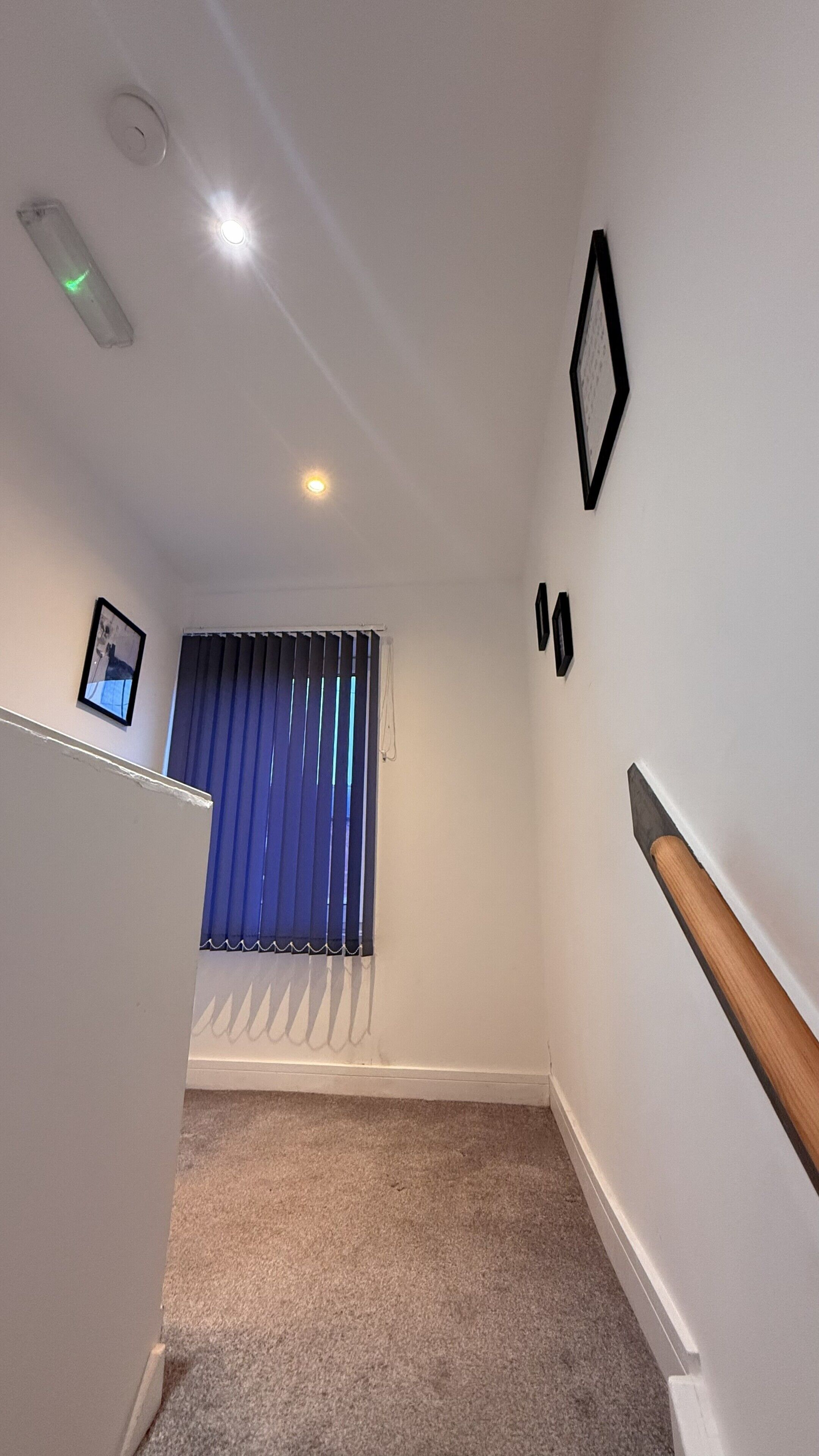 A narrow hallway with a carpeted floor, a window with blue blinds, a picture frame on the wall, and a wooden handrail.