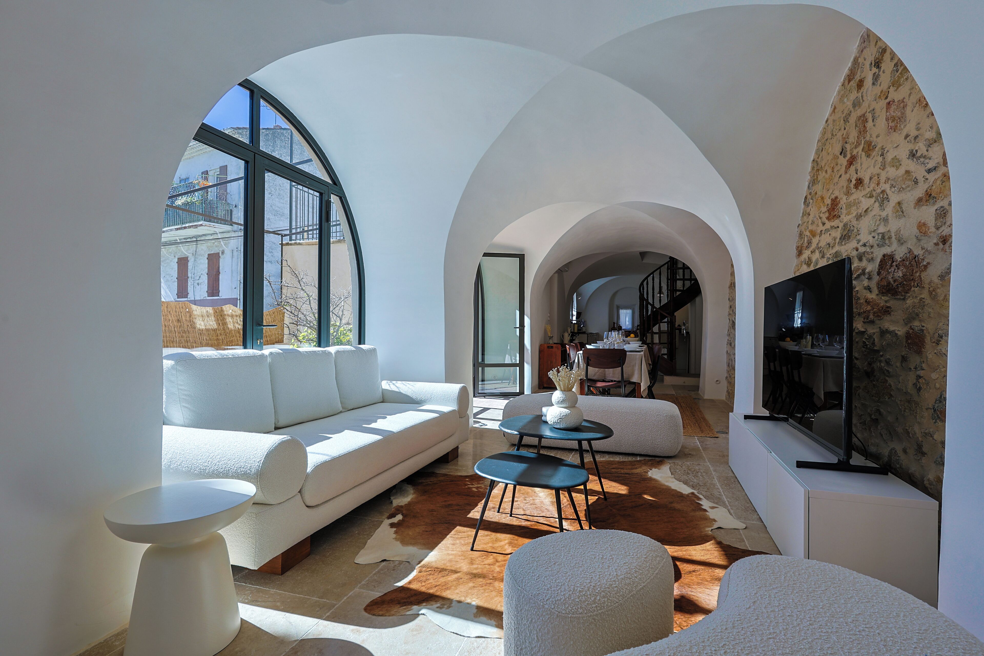 A modern living room with a white sofa, a round coffee table, and a television on a white cabinet. There is a large window providing a view of the outside.
