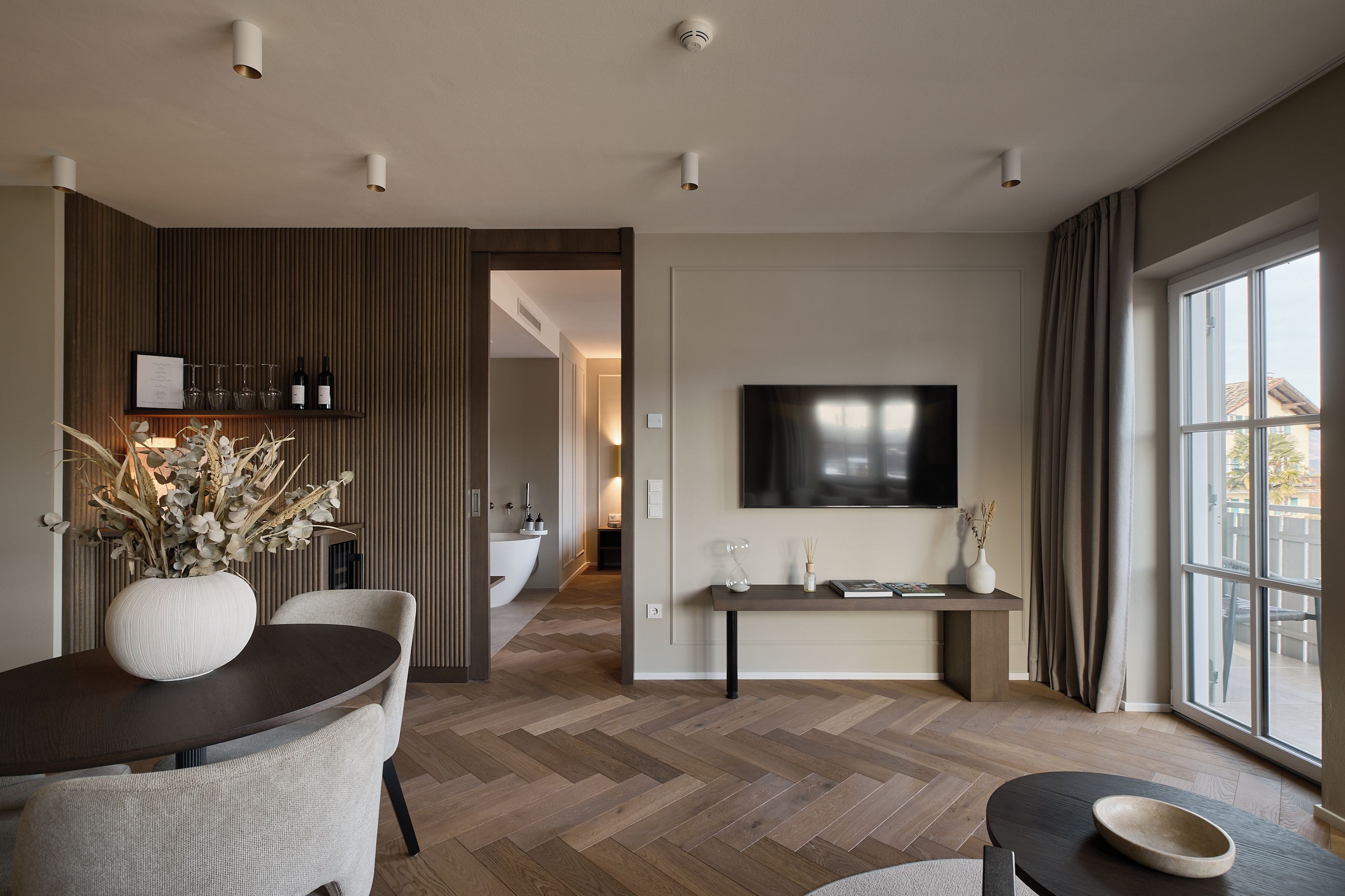 Ein modernes Wohnzimmer mit Essbereich, einem Wand-Fernseher und Blick durch große Fenster nach draußen.