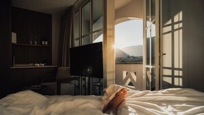A hotel room with a bed, a TV, a desk, and a view of the outdoors. - Lingers Suites & Residence (Appiano Sulla Strada del Vino)
