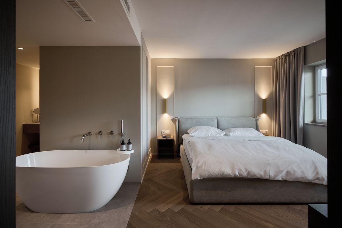 Una camera da letto moderna con una grande vasca da bagno freestanding, un letto con testiera grigia e lampade da comodino.