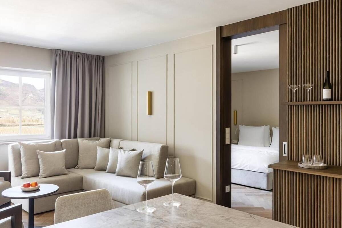 Een moderne hotelkamer met een sofa, een eettafel en een bed. Er staan twee wijnglazen op de tafel en een fles op het bijzettafeltje.