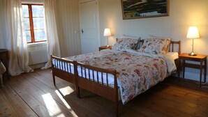 A bedroom with a wooden bed, floral bedding, two bedside tables with lamps, a window with curtains, and a painting on the wall. - Spacious Farmhouse – Skiing, Forest & Town. Pet-friendly! (Svenstavik)