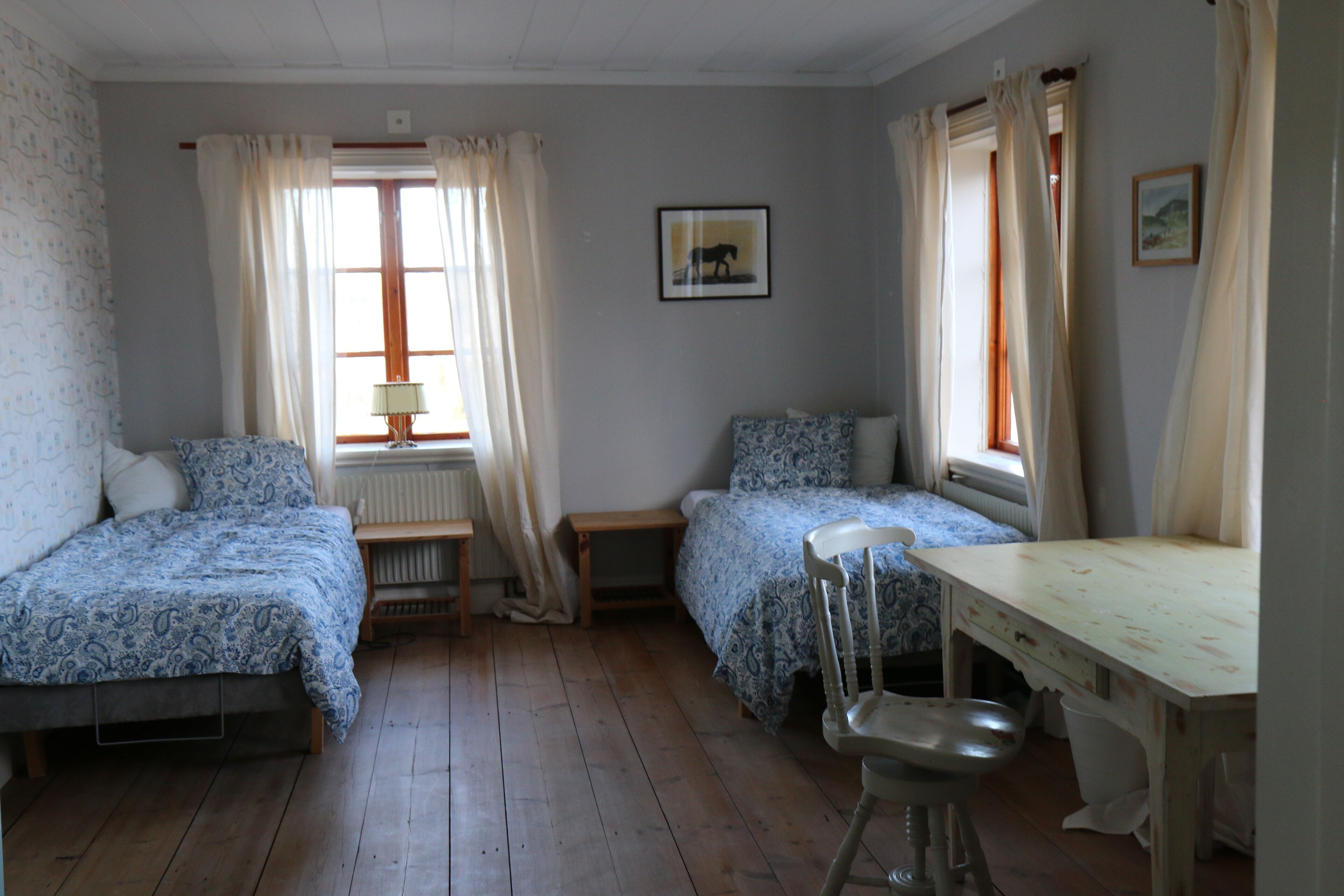 A room with two single beds, a wooden table, a chair, a radiator, and a window with curtains.