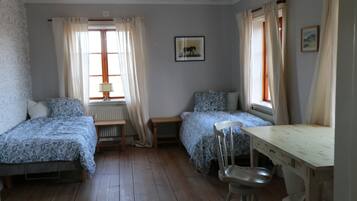 A room with two single beds, a wooden table, a chair, a radiator, and a window with curtains.