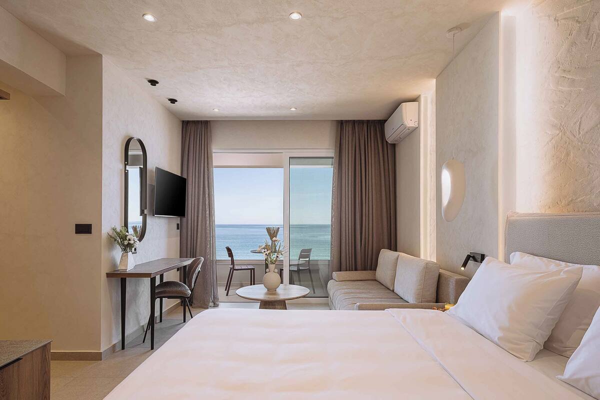 A hotel room with a large bed, a desk with a chair, a small table, a sofa, a TV mounted on the wall, and a view of the sea through the window.