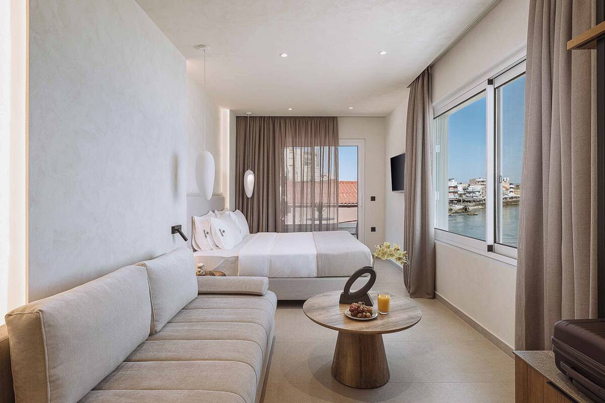 A hotel room with a sofa, a bed, a round wooden table with fruit and juice, and a view of the sea.