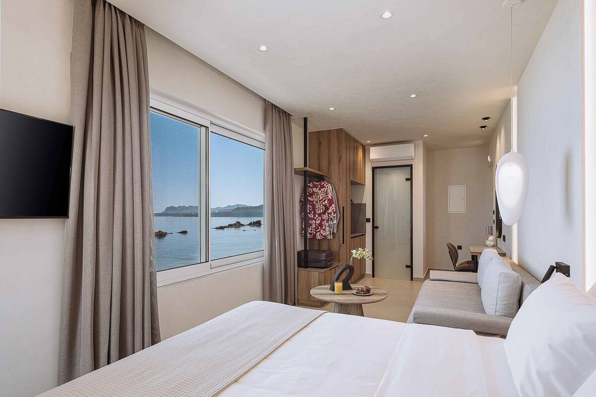A hotel room with a large bed, a smaller bed, a desk with a chair, a TV, and a view of the sea.