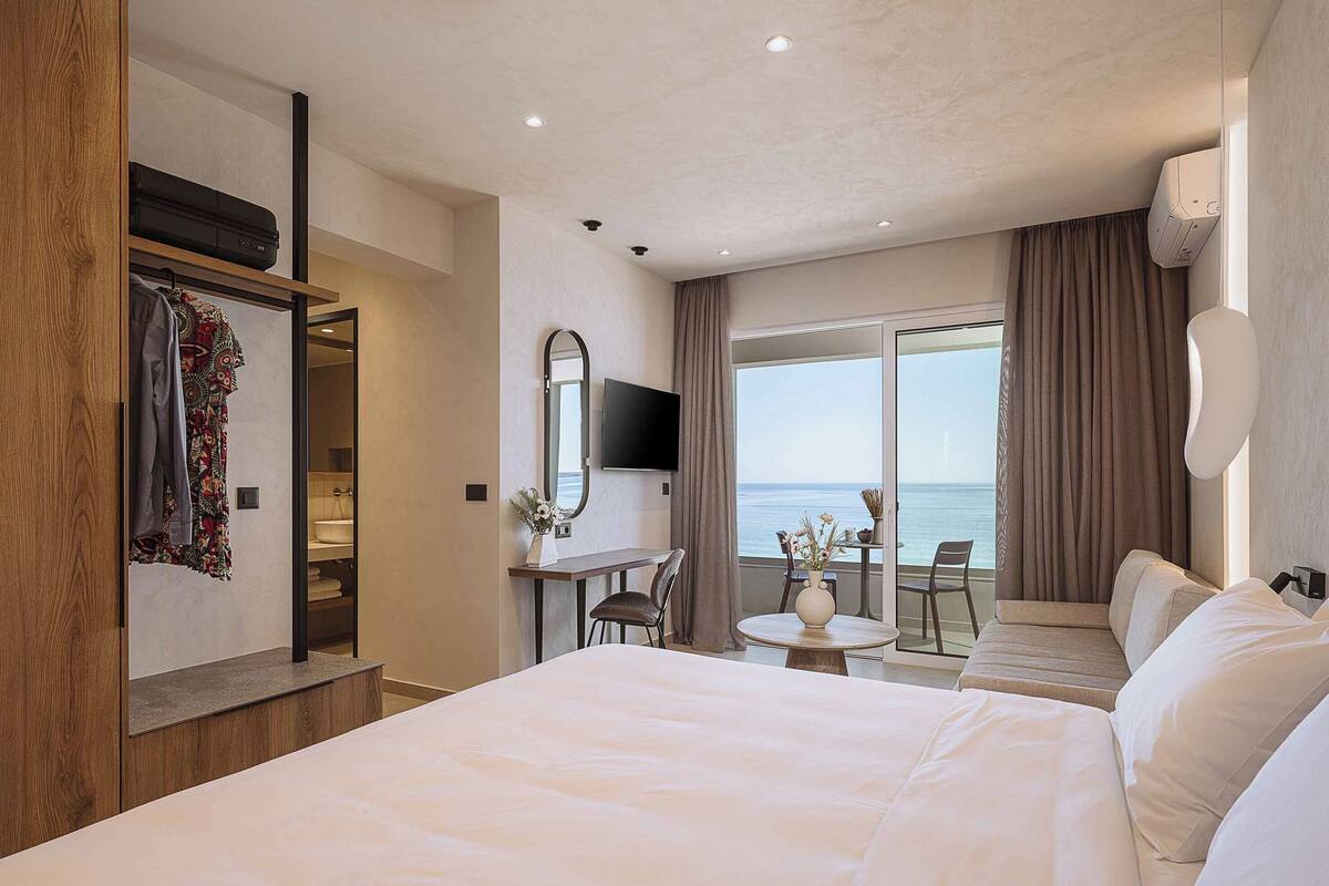 A hotel room with a large bed, a desk with a chair, a TV, and a view of the sea.