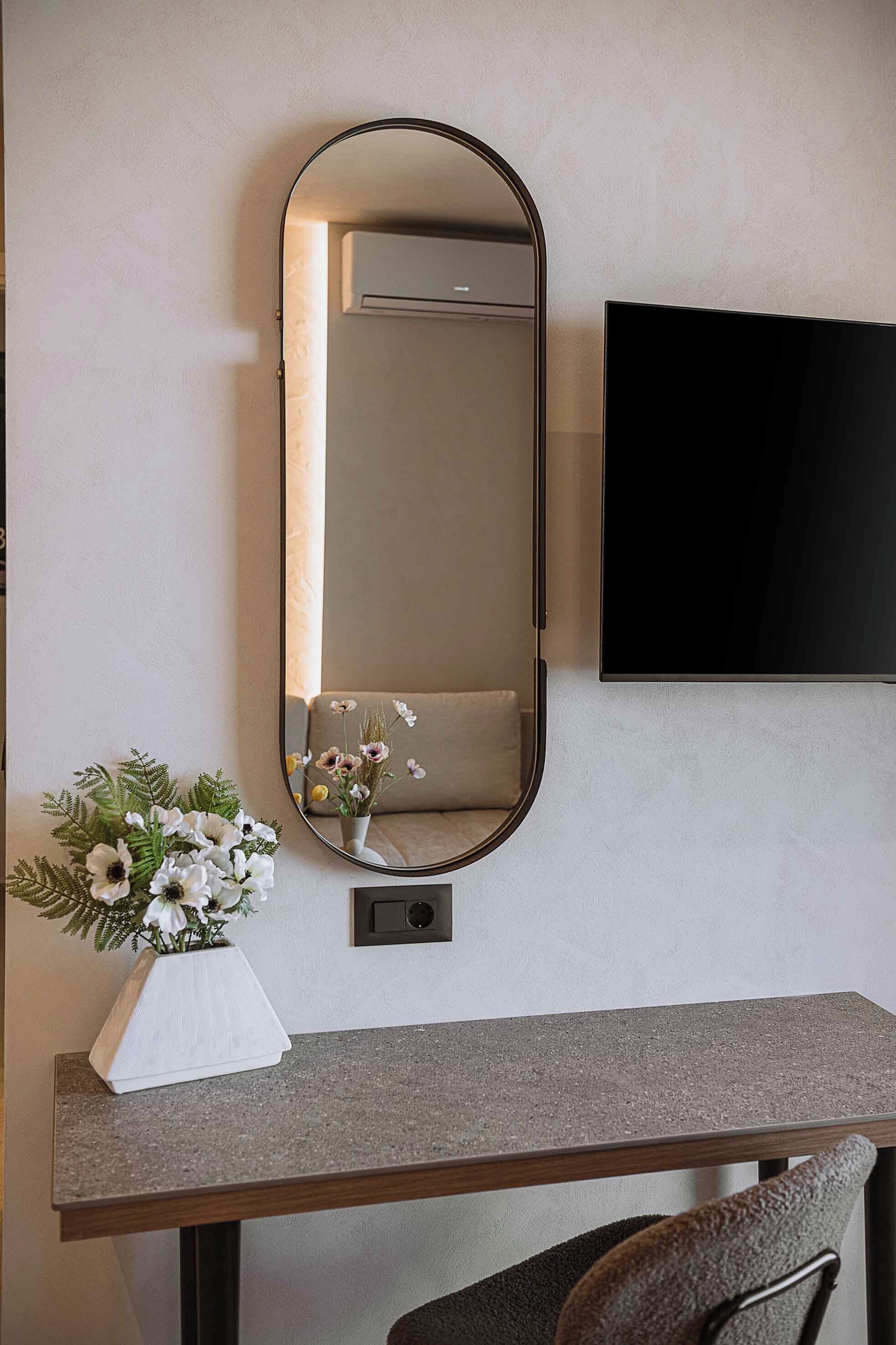 A room with a wall-mounted TV, a mirror, a small table with a vase of flowers, and a chair.