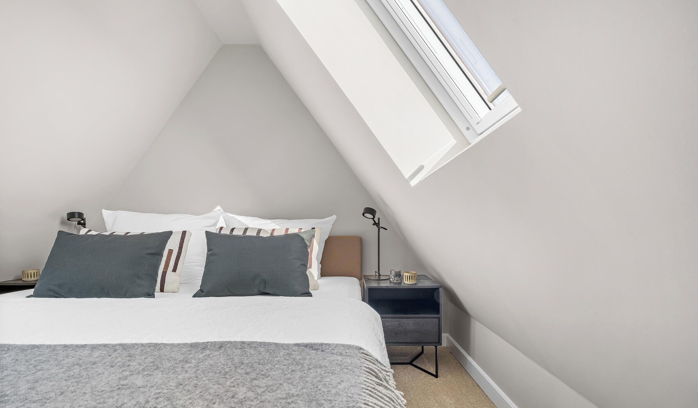 Ein Schlafzimmer mit schrägen Wänden, einem Bett mit vielen Kissen, einem Nachttisch mit Lampe und einem Fenster mit Klimaanlage.