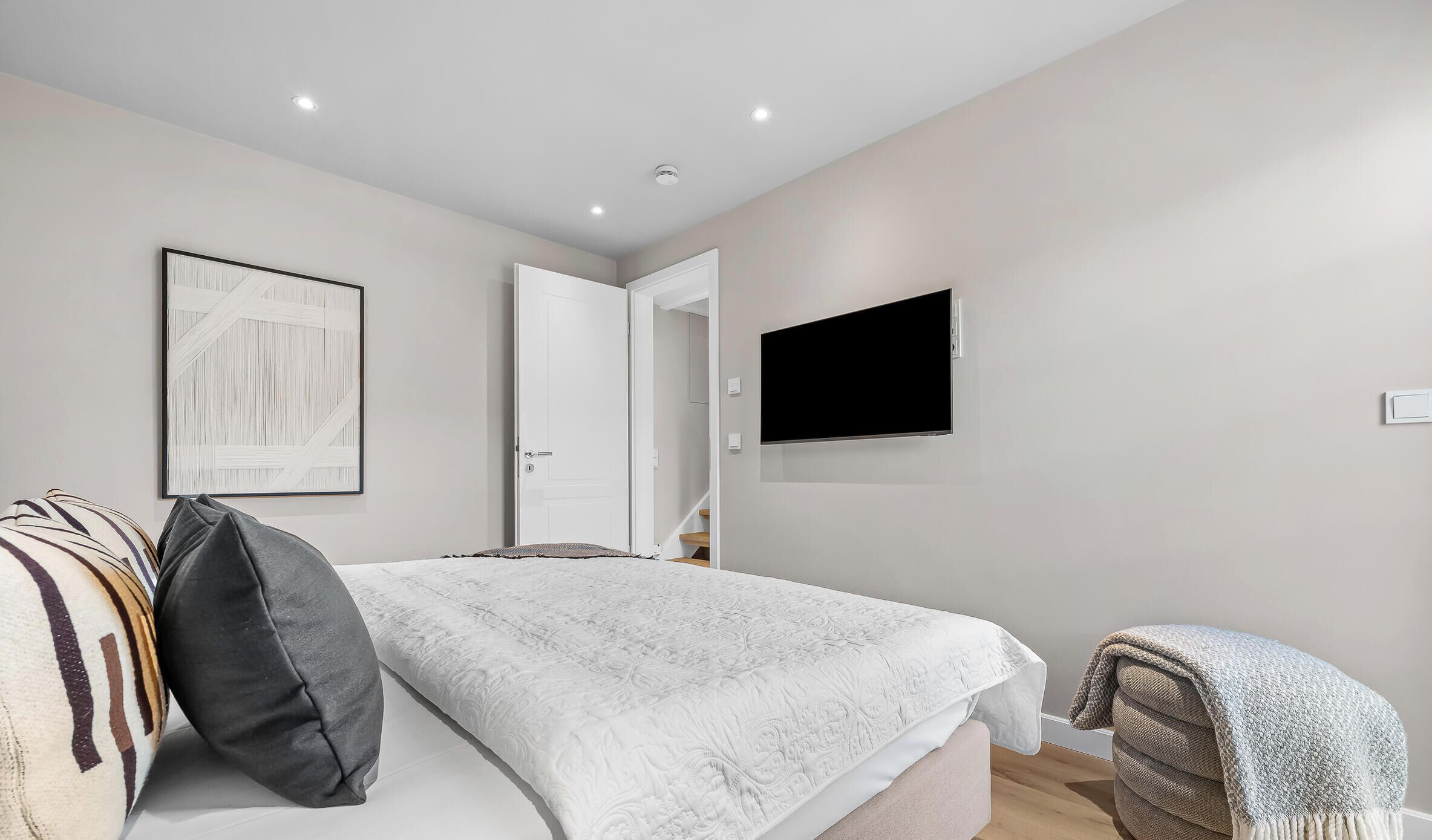 Une chambre à coucher avec un lit, un téléviseur fixé au mur, une porte donnant sur une autre pièce et un tableau encadré accroché au mur.