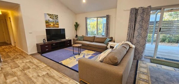 A living room with a sofa, a TV, a coffee table, and a large window with curtains. - The Long Jetty Beach House (Port Welshpool)