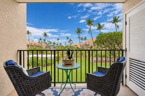 A balcony with two wicker chairs and a small table, overlooking a well-maintained lawn and palm trees. - Kamaole Sands 5-210 - Stunning 1BR/2BA, King Bed, Center-Cut Bldg 5, AC, Wi-Fi (Kihei)