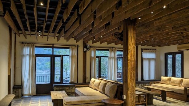 A cozy room with a stone floor, wooden ceiling beams, and large windows. There are two sofas and a bench. - Landmark Farmhouse (Bharatpur)