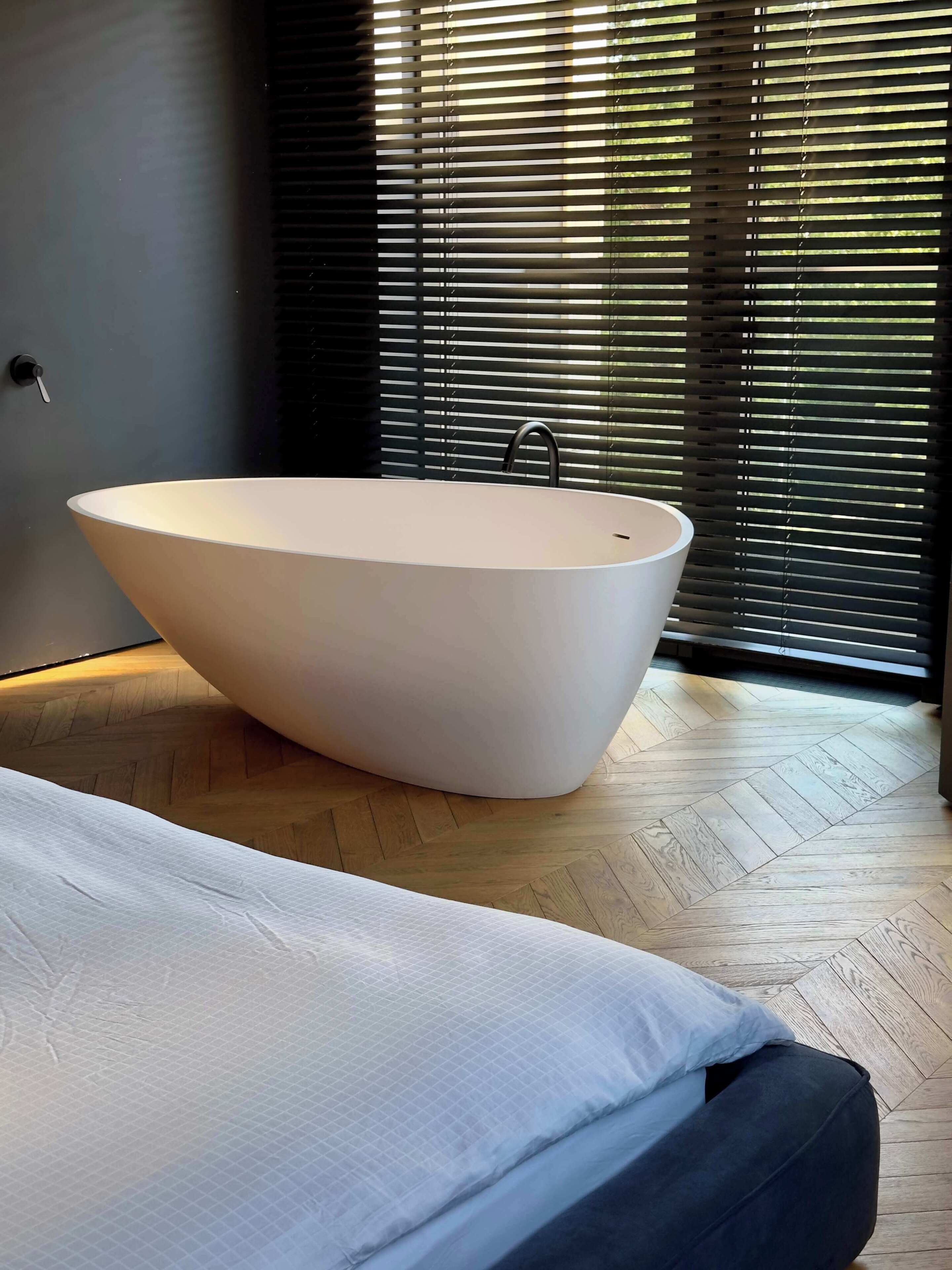 A modern bathroom with a freestanding bathtub, a wooden herringbone floor, and a large window with blinds.