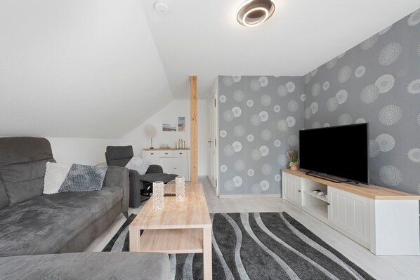 A modern living room with a grey sofa, a wooden coffee table, a TV on a white cabinet, and patterned wallpaper.