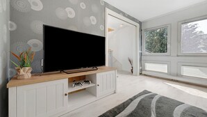 A modern living room with a flat-screen TV on a wooden console, a grey patterned wall, and large windows.