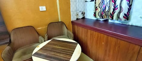 Um quarto pequeno com duas cadeiras de couro marrom, uma mesa redonda de madeira, um balcão de madeira e uma cortina estampada.