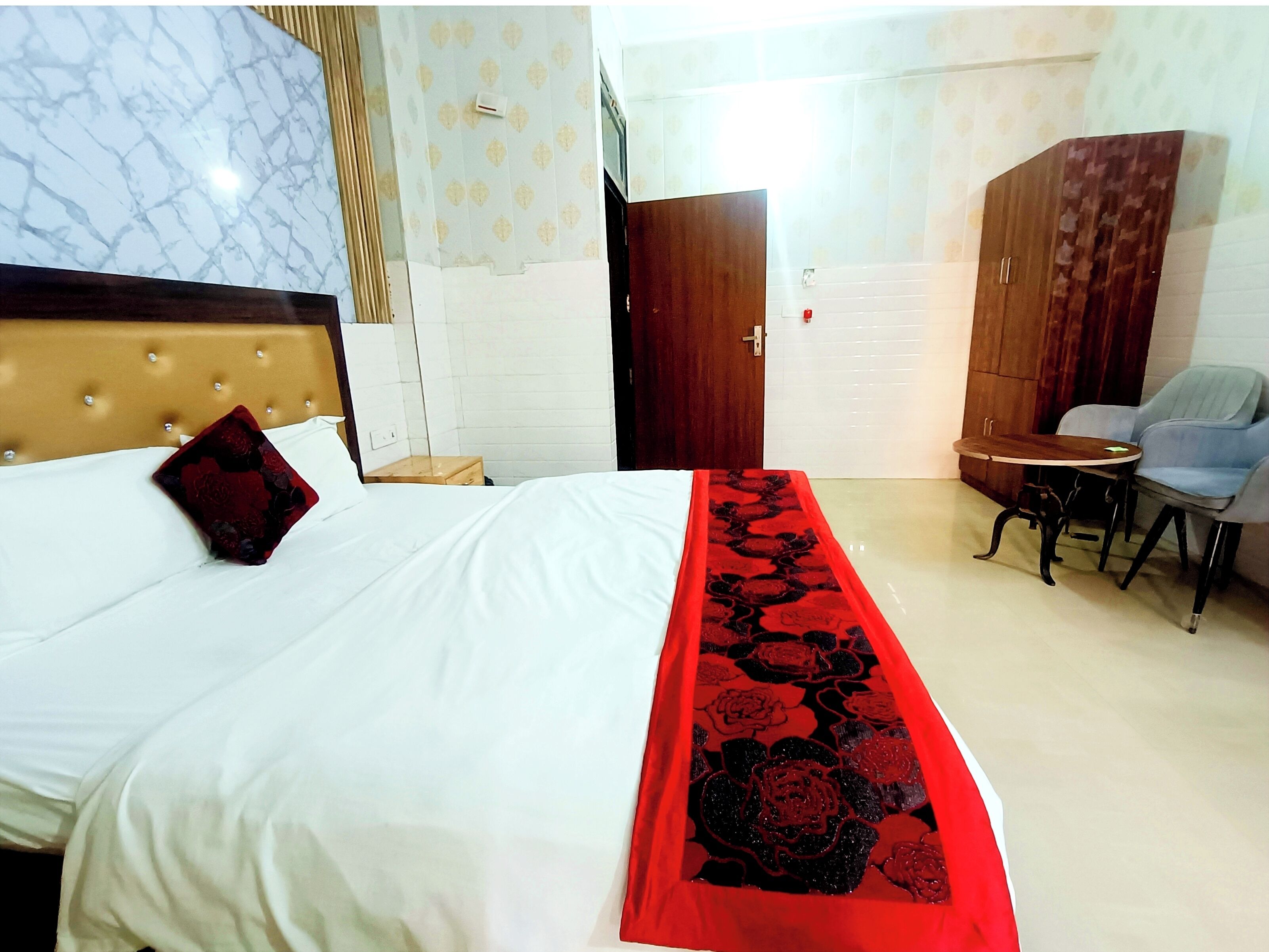A hotel room with a bed, a wooden headboard, a red floral bedspread, a small round table with two chairs, and a wardrobe.