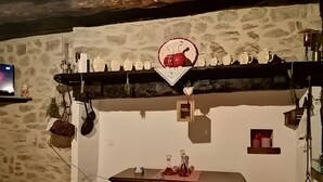 Una habitación rústica con pared de piedra, una mesa de madera con un adorno, y una estantería con diversos utensilios de cocina.