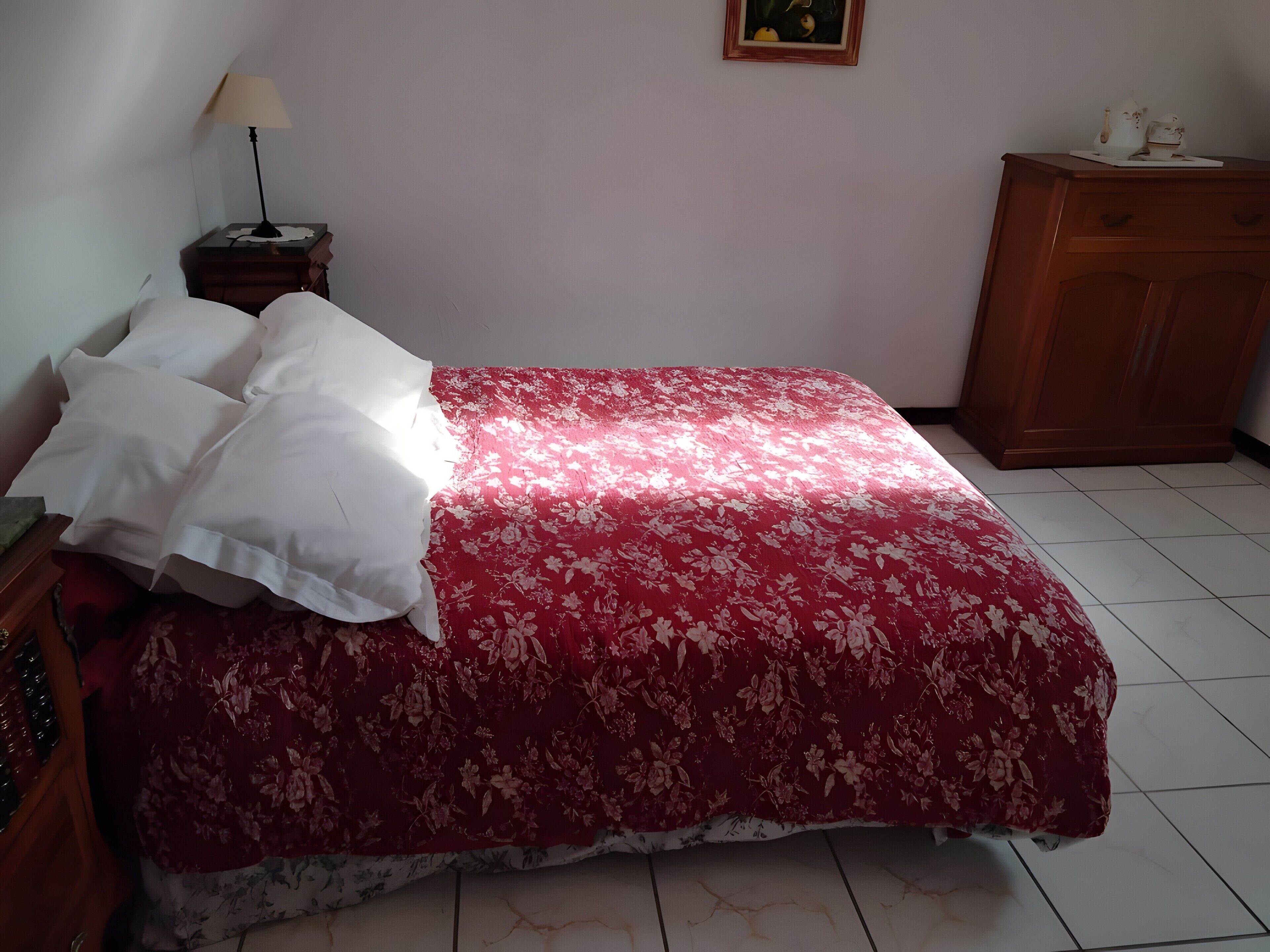 A bed with a floral bedspread, a nightstand with a lamp, a wooden dresser, and a framed picture on the wall.