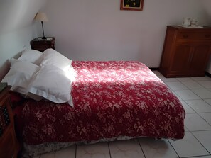 Een bed met een bloemenprint sprei, een nachtkastje met een lamp, een houten dressoir en een lijst met een foto aan de muur.