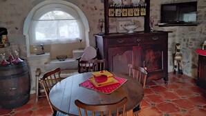 Un comedor rústico con una mesa redonda de madera, sillas, un aparador de madera con estantes, un televisor montado en la pared y un ventanal con arco decorativo.