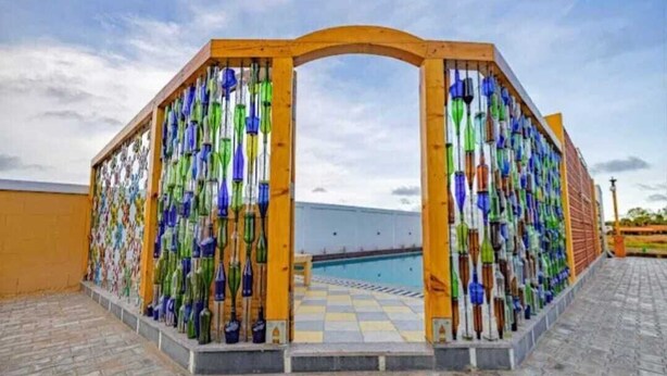 Un espace extérieur abrité doté d’une piscine, entouré d’une structure en bois agrémentée de bouteilles en verre aux couleurs vives.