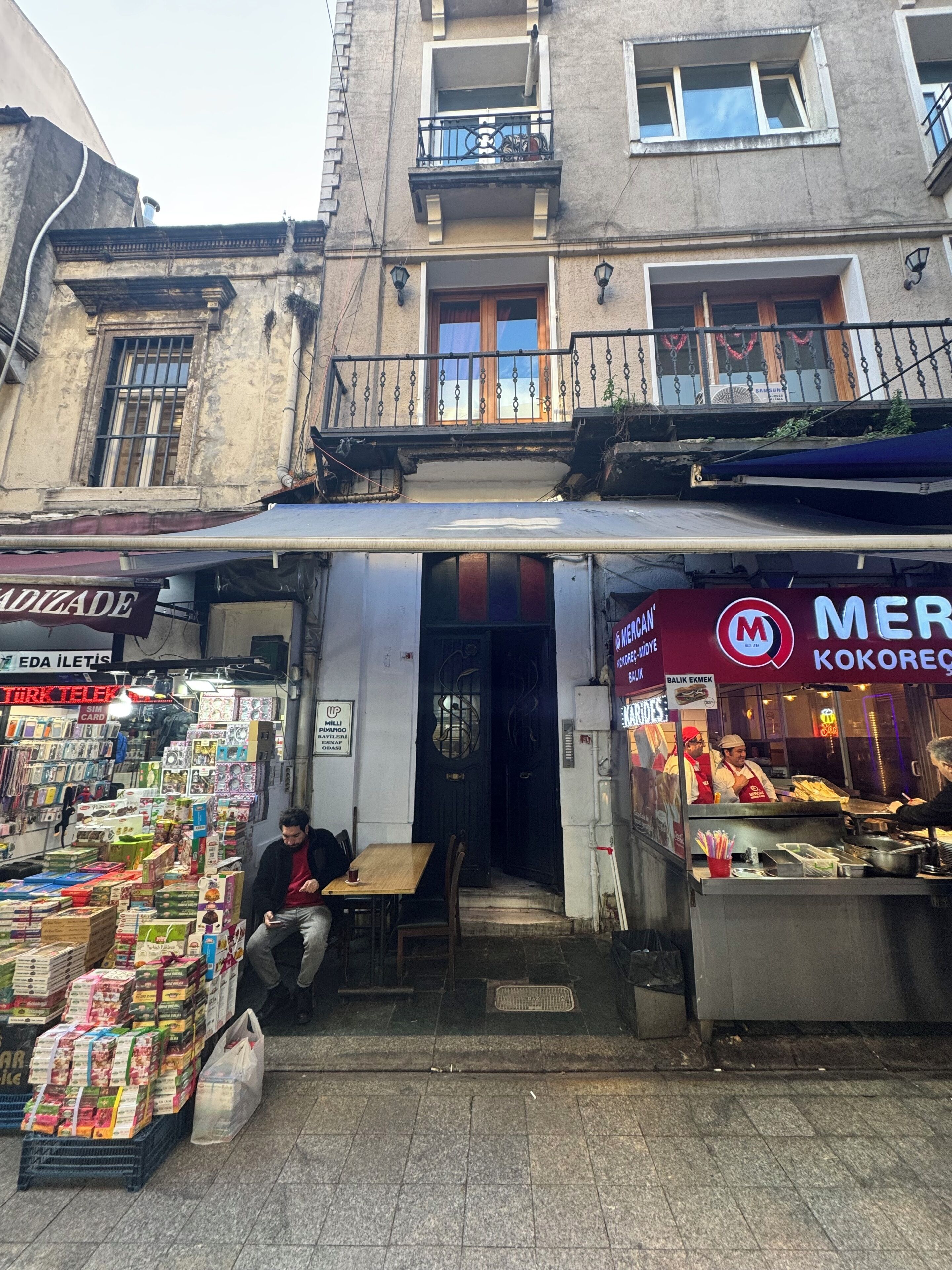 A street view with a shop selling various goods, a food stall, and a building with a balcony.