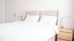 Una cama bien hecha con sábanas blancas, una mesita de noche con una lámpara y una puerta cerrada.