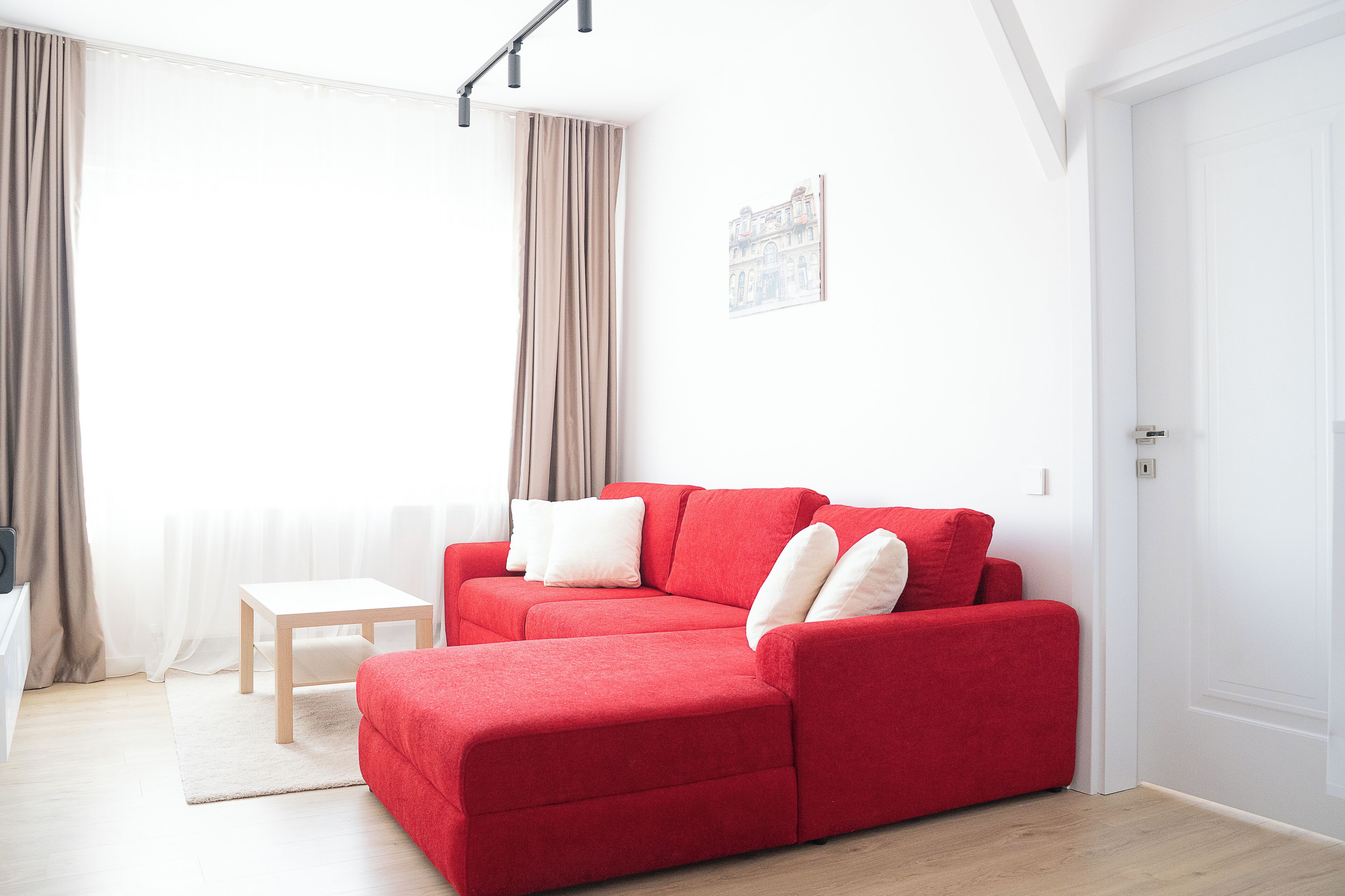 A red sofa with white cushions, a small wooden table, and a white rug in a bright room with white walls and a white door.