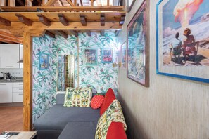 Une pièce chaleureuse avec un plafond en bois, un canapé gris agrémenté de coussins à motifs, un mur décoré de tableaux encadrés et, au fond, une cuisine.