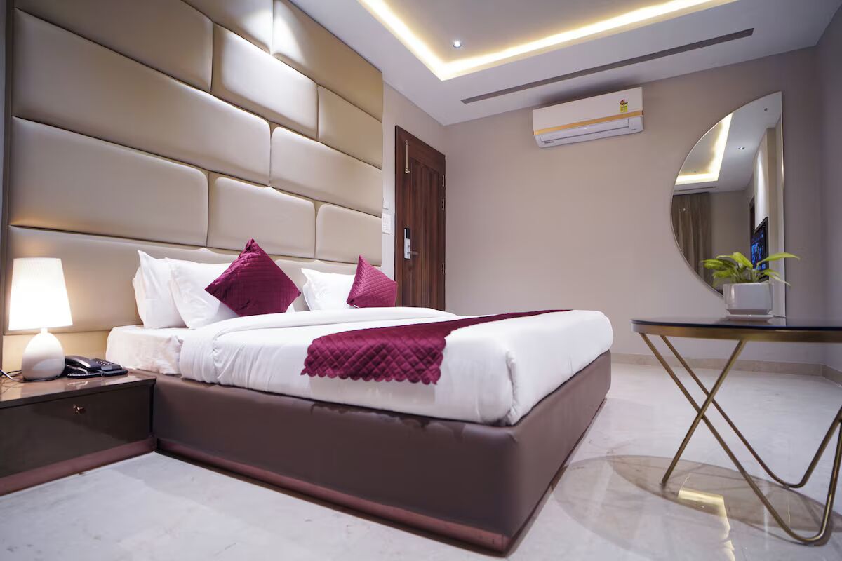 A modern hotel room with a large bed, a bedside table with a lamp, a small table with a plant, and a mirror on the wall.