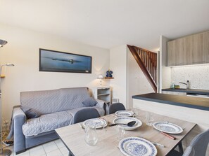 Un salon moderne avec un canapé gris, une table à manger dressée avec de l’assiette et des couverts, une cuisine avec des meubles en bois et un escalier.