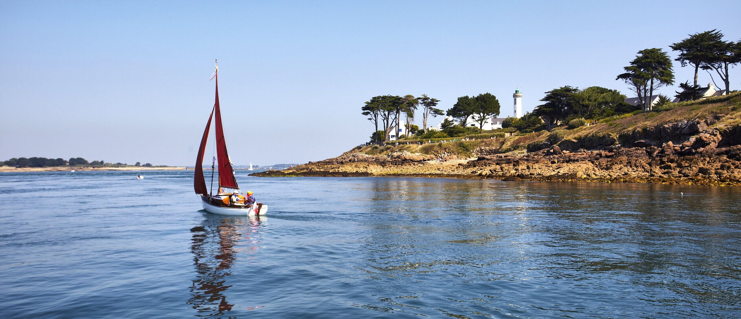 A sailboat on calm water with a rocky shoreline and trees in the background.