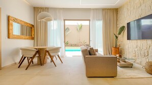 A modern living room with a sofa, a dining table with chairs, a TV mounted on the wall, and a view of the outdoor pool area. - Bungalow Medina - Heated swimming pool not overlooked (Marrakech)