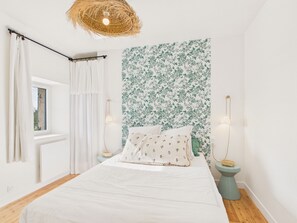 A bedroom with a bed, white curtains, a window, a wall with floral wallpaper, and two bedside lamps. - Beach 300m away, house for 7, garden and veranda (Plouguerneau)
