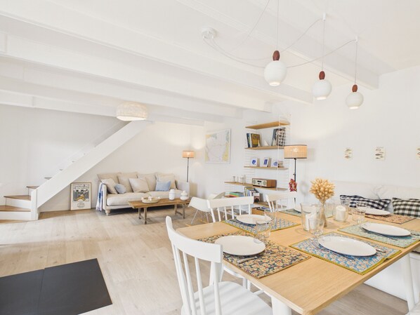 A spacious living area with a dining table set for four, a sofa, and a staircase leading to an upper level. - Beach 300m away, house for 7, garden and veranda (Plouguerneau)