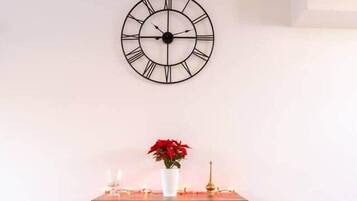 A room with a wooden floor, a wall-mounted clock, a small wooden table with a vase of red flowers, and a candle holder.