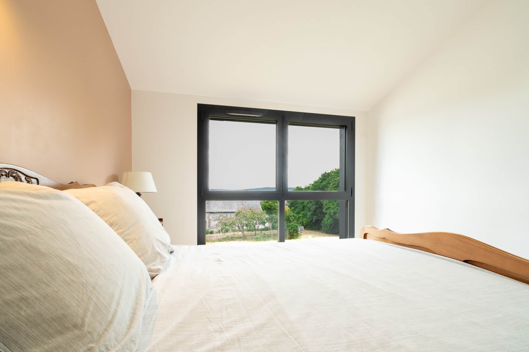 Une chambre avec une grande fenêtre, un lit avec une literie blanche et une tête de lit en bois.