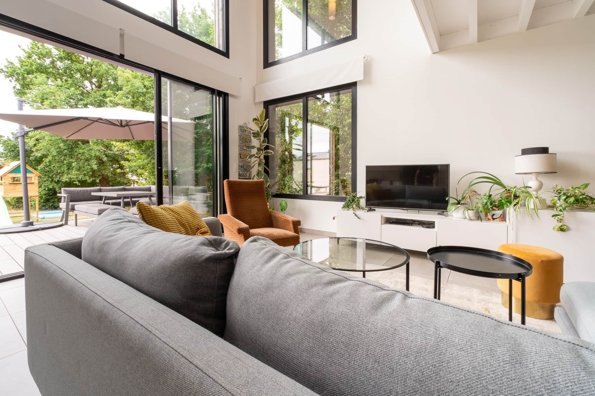 A modern living room with a large gray sofa, a brown armchair, a glass coffee table, a TV, and a white console table with plants.