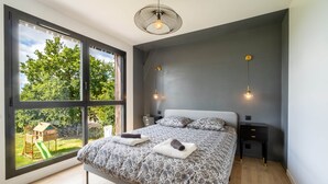 A bedroom with a large window, a bed with patterned bedding, bedside tables, and a hanging light fixture. - Architect-designed house in the heart of nature (Guichen)