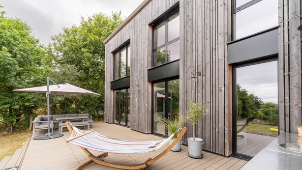 A modern house with a wooden deck, a hammock, outdoor furniture, and a large window. - Architect-designed house in the heart of nature (Guichen)